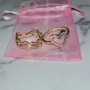 Guldplaterade ringar med hjärtdetaljer - Två snygga guldplaterade ringar, en med öppet hjärta och en med flera sammankopplade hjärtan runt om. Perfekta för att ge din look en söt och trendig touch. Kommer i en rosa organzapåse.