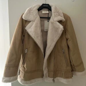 Beige teddyjacka från Pull&Bear - Snygg beige teddyjacka från Pull&Bear med mjukt vitt foder och krage. Jackan har dragkedja, två fickor med dragkedjor och bältdetaljer vid nederkanten. Perfekt för kalla dagar med sin mysiga och trendiga look.