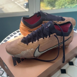 Nike Air Zoom Mercurial Vapor 15 ”Generation Pack” - Snygga Nike Air Zoom Mercurial Vapor 15 Generation Pack fotbollsskor i guld-brons med svarta detaljer och rosa innersula som var VM 2022 skorna. Ovandel i syntetmaterial med svart snörning och stor silverfärgad Swoosh. Yttersulan har metalliska och rosa detaljer för grymt grepp på planen. Väldigt fint skick, skorna är size 40, köpta från unisport och har även kvittot, box och påse tillkommer, Ny pris på unisport är 2949, säljer för 1699. Vid frågor eller funderingar hör bara av er! 