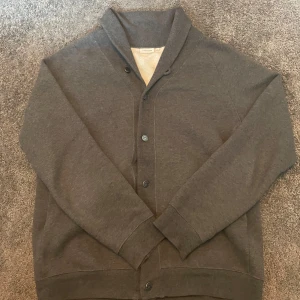 Grå kofta från J.Lindeberg - Mörkgrå kofta från J.Lindeberg i storlek L. Koftan har en snygg sjalkrage, knäppning med knappar framtill och långa ärmar. Hör av er vid frågor