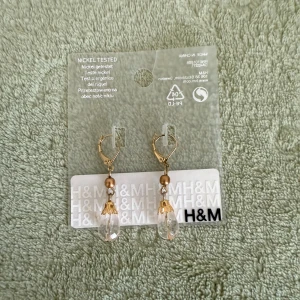 Guldiga örhängen från H&M - Snygga örhängen från H&M med guldfärgade detaljer och droppformade, klara pärlor. Örhängena har en liten rund pärla upptill och dekorativa guldfärgade hållare kring den genomskinliga droppen. Perfekta för att lyfta din look med lite extra bling. 