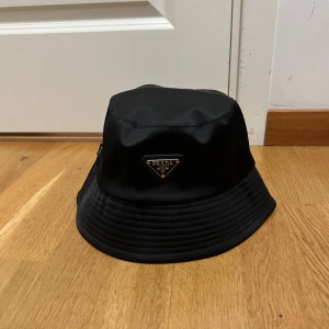 Svart bucket hat från Prada - Stilren svart bucket hat från Prada med klassisk siluett och ikonisk metallogo framtill. Hatten är tillverkad i glansig syntet och har bred, nedåtlutande brätte. Perfekt accessoar för att lyfta din outfit med en lyxig touch.
