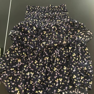 Blommig kortklänning i mörkblått - Supersöt kortklänning i mörkblått med små blommor i gult, lila och rosa. Klänningen har smock upptill och volangig kjol, perfekt för sommarens alla tillfällen. Lätt och luftig, med korta ärmar och skön passform.