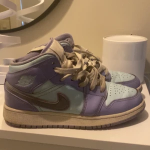 Nike Air Jordan 1 i lila och mint - Snygga Nike Air Jordan 1 sneakers i en unik färgkombination av lila och mintgrönt med beige detaljer och snörning. Skorna har klassisk hög siluett, rund tå och ikonisk Swoosh-logga på sidan. Perfekta för dig som vill sticka ut med din stil.