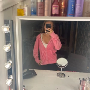 Juicy Couture rosa velourjacka  - Juicy Couture hoodie i rosa velour med dragkedja och huva. Ikoniskt guldtryck med palmer och text på ryggen. Mjuk och skön, perfekt för en chill look. Klassisk modell med långa ärmar och fickor framtill.