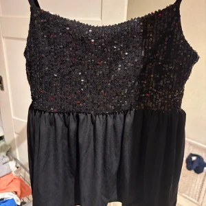 Svart paljettopp från Gina Tricot - Supersnygg svart topp från Gina Tricot med smala axelband och glittriga paljetter över hela överdelen. Nedre delen är i mjukt tyg som faller snyggt. Perfekt för dig som vill sticka ut med lite extra bling!