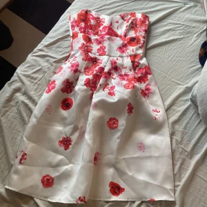 Vit blommig klänning från Goddiva - Superfin axelbandslös klänning från Goddiva London i vit satin med röda och rosa blommor. Klänningen har en figurnära överdel, utställd kjol och dragkedja bak. Perfekt för sommarens alla tillfällen när du vill sticka ut lite extra.