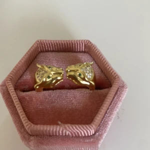 Guldfärgad ring med leopardhuvuden - Cool ring i guldfärg med två leopardhuvuden som möts framtill. Huvudena har små gröna stenar som ögon och glittrande stenar på sidorna, vilket ger en lyxig och unik look. Perfekt statement piece för dig som gillar djurmotiv och detaljer.