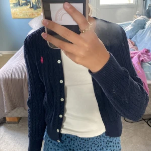 Mörkblå kofta från Polo Ralph Lauren - Mörkblå kabelstickad kofta från Polo Ralph Lauren med klassisk passform. Tröjan har vita knappar framtill och en rosa broderad logga på bröstet. Perfekt för lager-på-lager och snygg till både jeans och kjol. 