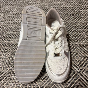 Vita sneakers med silvriga detaljer - Snygga vita sneakers från Inci med silvriga detaljer och mönstrad textur på sidorna. Skorna har snörning och en platt sula, perfekta för en sportig och trendig look. Passar till många olika outfits och ger en fräsch känsla.