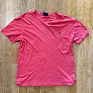 Rosa t-shirt från Polo Ralph Lauren - Snygg rosa t-shirt från Polo Ralph Lauren med klassisk v-ringning och broderad marinblå logga på bröstet. Tillverkad i mjuk bomull och har korta ärmar. Perfekt för dig som gillar stilrena och bekväma plagg.