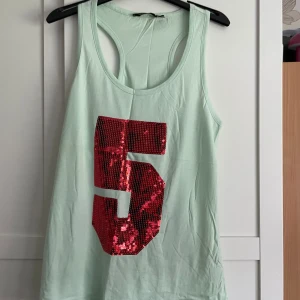 Mintgrön linne med paljettfemma - Snygg mintgrön linnetopp med stort rött paljett-nummer 5 framtill. Modellen är ärmlös med brottarrygg och tillverkad i mjuk bomull. Perfekt för dig som gillar sportig och glittrig stil.