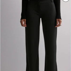 Svarta kostymbyxor - Snygga svarta kostymbyxor från Nelly med raka, lite vidare ben och låg midja. Byxorna har diskret knapp och dragkedja framtill samt hällor för bälte. Perfekta för en clean och stilren look.