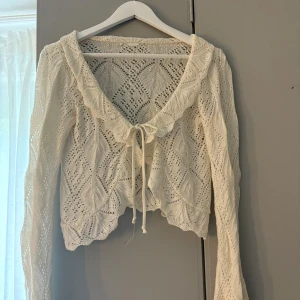 Vit stickad kofta med knyt och volang - Superfin vit stickad kofta med vackert hålmönster, volangkant och knyt framtill. Croppad modell med långa ärmar och romantisk känsla. Perfekt att slänga över en topp för en söt look. Köpt i Spanien, använd många enstaka gång