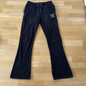 Juicy Couture svarta mjukisbyxor - Svarta mjukisbyxor från Juicy Couture i mjuk velour med bootcut-ben. Byxorna har snörning i midjan, två bakfickor och ett glittrigt JC-logo i strass på ena höften. Super sköna att ha hemma i soffan eller också ute. Perfekta för en chill och trendig look. Köparen står för frakt