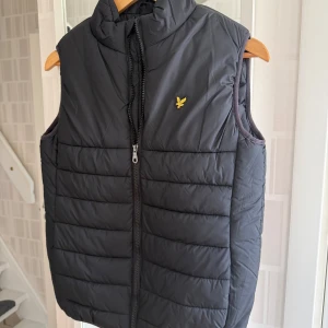 Svart dunväst från Lyle & Scott - Helt ny, dvs oanvänd snygg svart dunväst från Lyle & Scott Junior med gul logga på bröstet. Västen har hög krage, quiltad design och dragkedja framtill. Perfekt att slänga över en hoodie för en chill look. Storleken motsvarar ca 170 cl.