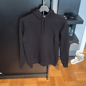 Svart stickad hoodie från Dressmann - Svart stickad hoodie från Dressmann i storlek S. Tröjan har huva med snörning och långa ärmar. Ribbstickade muddar vid ärmslut och nederkant ger en clean look. Perfekt för dig som gillar enkel och stilren streetwear.