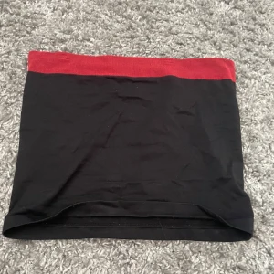 Svart kortkjol med röd midja - Enkel och stilren svart kortkjol med bred röd midjeresår. Kjolen har en tight passform och är perfekt för dig som gillar minimalistisk stil med en färgklick. Passar snyggt till både sneakers och boots.