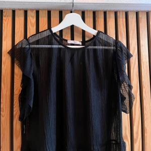 Svart transparent blus från MNG - Säljer en svart transparent blus från MNG med volangdetaljer på ärmarna. Blusen är gjord i ett lätt, plisserat syntetmaterial och har en luftig känsla. Perfekt för dig som gillar en trendig och edgy stil med extra volym på axlarna. Liten i storleken!