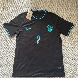 Svart Brasilien Vini Jr. Nike fotbollströja - Snygg svart Brasilien fotbollströja från Nike med Vini Jr. och nummer 7 i blått på ryggen. Tillverkad i lätt och ventilerande material, perfekt för match eller träning. Den är lite stor i storleken så om du är en liten M kan den passa