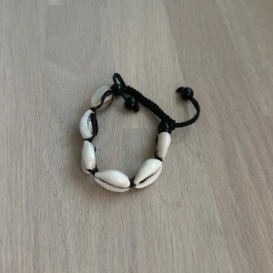 Svart snäckarmband med justerbar knytning - Snyggt armband med vita snäckor trädda på svart snöre och justerbar knytning. Perfekt accessoar för en chill sommarlook eller strandhäng. Snäckorna är ovala och ger en naturlig vibe, medan de svarta detaljerna gör armbandet stilrent och enkelt att matcha.