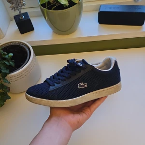 Lacoste sneakers - Snygga marinblå sneakers från Lacoste med mesh-yttermaterial och klassisk krokodil-logga på sidan. Skorna har vita detaljer vid häl och innersula, samt platt sula och rund tå. Perfekta för en stilren och sportig look. Storlek 38