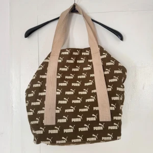 Brun väska från Puma - Cool brun shoppingväska från Puma med beigea handtag och all-over logomönster. Väskan är rymlig och har innerficka med dragkedja. Perfekt för att bära med sig allt man behöver till skolan eller stan.