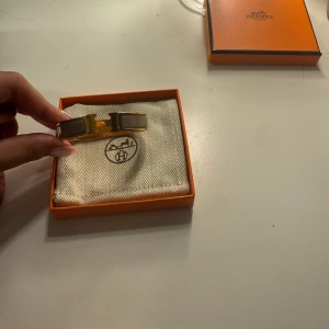 Hermès guldarmband med beige detaljer - Snyggt armband från Hermès i guld med beige/grå färgl. Kommer i original ask och tygpåse. Inköpt för 8300kr. Den är lite böjd, men inget som påverkar funktionen!