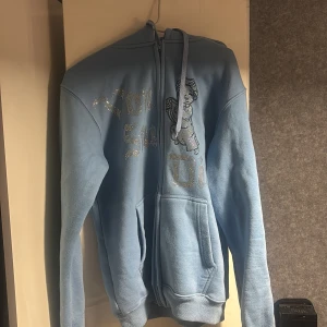 Ljusblå vonstudio Hoodie - Ljusblå hoodie med dragkedja och huva. Framsidan har ett glittrigt tryck med text och en ängel. Perfekt för dig som gillar en chill och snygg stil med lite extra bling.