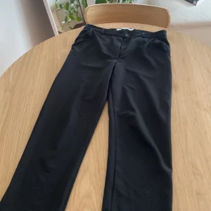 Svarta kostymbyxor från 157, stl S - Klassiska svarta kostymbyxor från 157 med raka ben och låg midja. Byxorna har knapp och dragkedja framtill och är perfekta för en clean och stilren look. Materialet känns mjukt och har en lätt stretch.