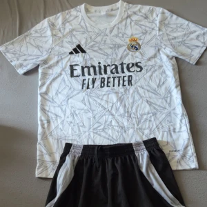 Real Madrid vit tränings ser Adidas - Snygg vit Real Madrid fotbolls set från Adidas med grått grafiskt mönster och klubbmärke på bröstet. Tröjan har svart Adidas-logga och sponsortryck framtill. Tillverkad i lätt och ventilerande material, perfekt för träning eller match.