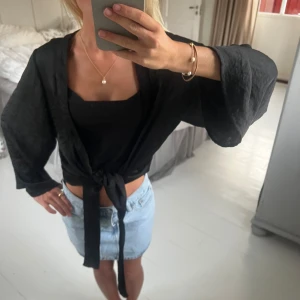 Svart silkes kofta/blus - Säljer en svart silkes kofta som man kan styla som en blus eller hur man vill. Vida ärmar och knytband framtill. glansig yta. Perfekt att slänga över ett linne eller topp. Ginatricot 
