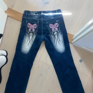 Crazy Age jeans - Unika blå jeans från Crazy Age med slitna detaljer och coolt rosa broderi på fickorna och framsidan. Jeansen har vita slitningar och svarta broderade mönster 