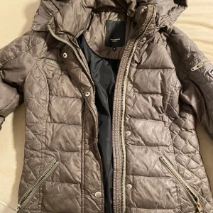 Brun quiltad dunjacka med huva - Snygg brun dunjacka från Rockandblue med quiltat mönster och dragkedjor i silver. Jackan har huva, flera fickor och en modern look. Perfekt för kalla dagar när du vill vara både varm och stilren.