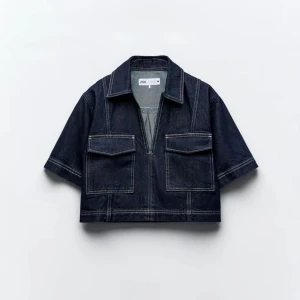 Mörkblå croppad jeansjacka från Zara - Snygg croppad jeansjacka från Zara i mörkblå denim med kontrastsömmar. Jackan har korta ärmar, två stora bröstfickor med lock och en halv dragkedja framtill. Perfekt för dig som vill ha en trendig och avslappnad look.