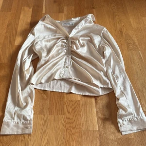 Beige omlottblus från Giracoo - Säljer en beige omlottblus från Giracoo i satinliknande material. Blusen har v-ringning, långa ärmar med knappar vid ärmslut och snygg rynkdetalj framtill med knappar längs hela framsidan. Perfekt för dig som gillar en chic och trendig look.