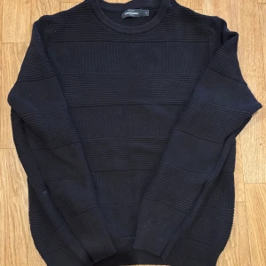 Svart ribbad stickad tröja Dressmann - Snygg svart stickad tröja från Dressmann i storlek M. Tröjan har breda ribbade partier som ger en cool struktur och rund halsringning. Perfekt för dig som gillar en stilren och enkel look med lite edge.