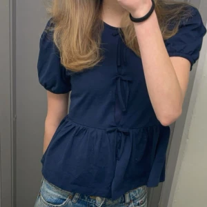 Mörkblå blus med knyt - Supersöt mörkblå blus med puffärmar och tre knyt framtill. Modellen har en lätt utställd peplum-nederdel och rund halsringning. Perfekt för dig som gillar en feminin och trendig look. Tunn och skön bomullskänsla.