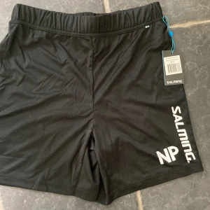 Svarta träningsshorts från Salming - Svarta sportiga shorts från Salming med vit logga på ena benet. De har elastisk midja och är tillverkade i ett lätt och smidigt syntetmaterial som passar perfekt för träning. Snygg minimalistisk design och bekväm passform. Felköp