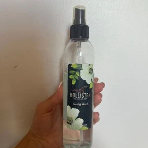Hollister Seacliff Beach body mist - Fräsch body mist från Hollister i doften Seacliff Beach. Flaskan är genomskinlig med svart spraykork och har en snygg etikett med vita blommor och gröna blad. Perfekt för dig som gillar lätta, somriga dofter och vill ha en enkel sprayflaska att ta med.