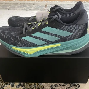 Adidas Supernova löparskor grå/turkos - Snygga Adidas Supernova sneakers vunna i en frågatävling av adidas. Skorna är i mörkgrått med turkosa och ljusgula detaljer. Ovandel i ventilerande syntetmaterial, klassiska tre ränder på sidan och Dreamstrike+ sula för extra komfort. Perfekta för löpning eller träning, med snörning och platt sula.