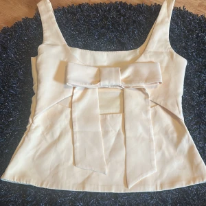 Beige linnetopp med stor rosett - Supersöt beige linnetopp med breda axelband och en stor rosett framtill som ger en extra touch. Toppen har en lätt figursydd passform och är gjord i ett slätt, mjukt material som känns lyxigt. Perfekt för dig som vill sticka ut med en feminin och trendig look.