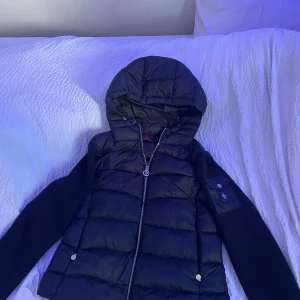 Svart Moncler dunjacka med huva - Säljer en svart Moncler dunjacka med huva och dragkedja. Jackan har quiltad front och rygg i glansigt material, samt stickade ärmar med Moncler-logga och ficka på ena ärmen. Perfekt för kyliga dagar och riktigt snygg streetstil.
