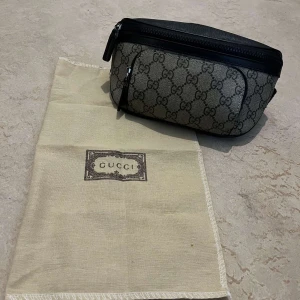 Gucci magväska i svart och beige - Säljer en snygg magväska från Gucci i beige och svart med klassiskt GG-mönster. Väskan har svarta skinn-detaljer, justerbart band och dragkedjor både fram och bak. Perfekt för att bära det viktigaste med stil.