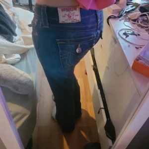 True Religion blå bootcut jeans - Säljer ett par blå jeans från True Religion med bootcut passform och snygga detaljerade bakfickor med knappar. Jeansen har låg midja och klassisk gul kontrastsöm. Materialet är jeans i bomull och de har en ikonisk True Religion patch bak. Ombedda till bootcut och är 76cm innerben och 36cm rätt över i midjan och 8cm gren pris går att diskutera vid snabb affär