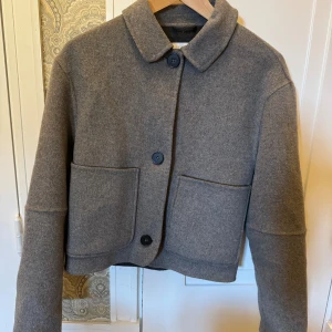 Grå ulljacka från Zara, croppad - Som Ny!!! Säljer en stilren grå croppad jacka från Zara i ullmix. Jackan har klassisk krage, stora framfickor och stängs med svarta knappar. Perfekt för dig som gillar minimalistisk och clean stil. Passar till både jeans och kjol. Köpare står för frakt eller så kan jag mötas upp nära Fältöversten/karlaplan