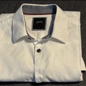 Vit skjorta från Burton London - Vit skjorta från Burton London med diskret paisleymönster i tyget. Klassisk krage, svarta knappar och kontrastfärgad insida på kragen. Långärmad och tillverkad i bomull, perfekt för en clean och stilren look.