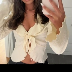 Krämvit blus med volang och knyt - Supersöt krämvit blus med volangkant längs urringningen och ärmsluten. Blusen har knyt framtill och lätt croppad passform. Tunt, flowigt tyg som ger en romantisk vibe och passar perfekt till jeans eller kjol.