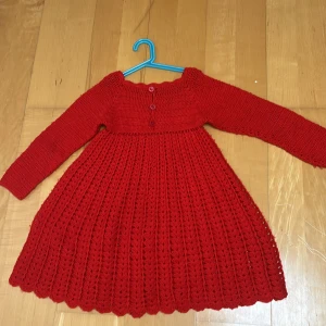 Röd stickad långklänning med knappar - Säljer en röd stickad långklänning med snyggt virkat mönster och tre knappar framtill. Klänningen har långärmade ärmar och en rundad hals med vågig kant. Perfekt för dig som gillar unika och handgjorda plagg med cozy vibes.