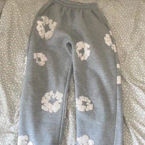 Grå mjukisbyxor Denim Tears med blommor - Säljer ett par grå mjukisbyxor från Denim Tears med vita blommiga tryck över hela byxorna. De har resår i midjan och är gjorda i mjukt bomullsmaterial. Perfekta för en chill och avslappnad stil.